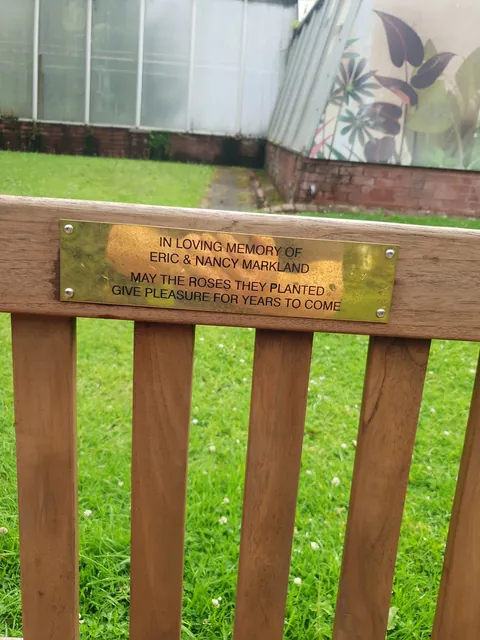 Polished brass memorial bench plaque for a couple, on a bench outside a park conservatory